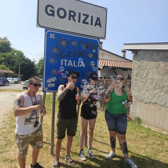 Vier Personen stehen vor einem Straßenschild für Gorizia, Italien. Sie pusten Seifenblasen und scheinen Spaß zu haben.