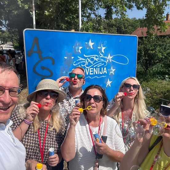 Fünf Erwachsene stehen vor einem blauen Schild mit Graffiti, lächeln und blasen Seifenblasen. Dahinter ist ein Gebäude mit einem roten Dach zu sehen.