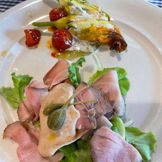 Ein weißer Teller zeigt ein Gericht mit Schinkenscheiben, Salat, Kirschtomaten und einer gebratenen Gemüsegarnitur. Das Gericht ist mit einer leichten Sauce beträufelt.