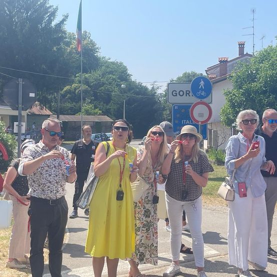 Eine Gruppe von Touristen in Sommerkleidung posiert auf einer Straße in Gorizia, Italien, mit einer Flagge und Schildern im Hintergrund.