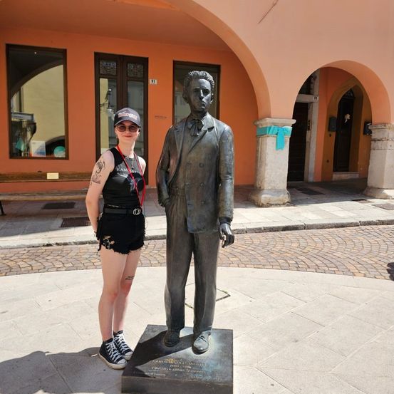 Eine Frau steht neben einer Statue eines Mannes in einem Anzug vor einem Gebäude mit Bogenfenstern. Die Statue ist mit einem blauen Band geschmückt.