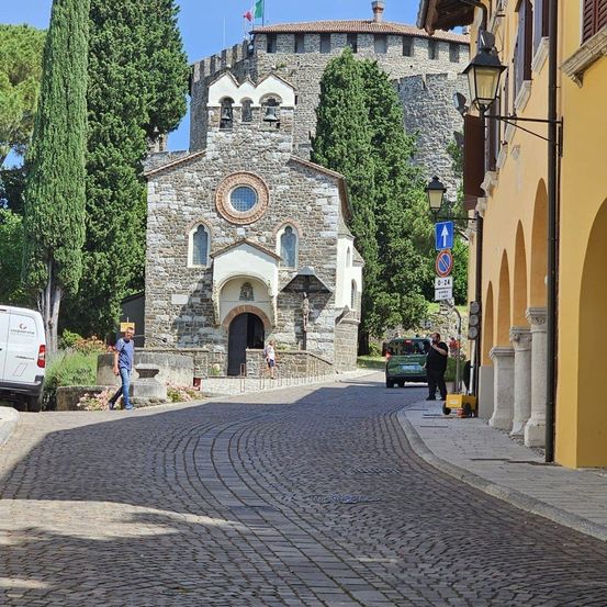 Eine Kopfsteinpflasterstraße mit einem weißen Lieferwagen auf der linken Seite. Eine Kirche mit Turm steht links. Ein grünes Auto fährt auf der Straße. Menschen gehen auf dem Bürgersteig.
