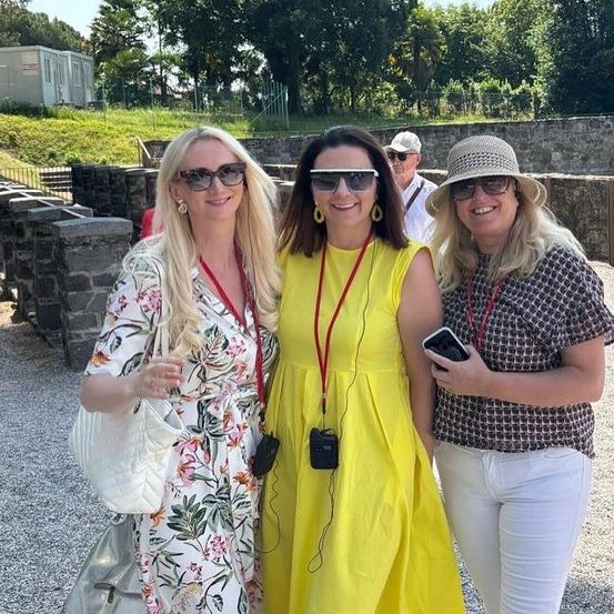 Drei Frauen stehen zusammen und lächeln für ein Foto. Die Frau auf der linken Seite trägt ein Blumenkleid und hält eine weiße Handtasche. Die Frau in der Mitte trägt ein gelbes Kleid mit einem roten Lanyard. Die Frau auf der rechten Seite trägt einen Hut und hält ein Telefon. Hinter ihnen befindet sich eine Steinmauer und ein Rasenbereich.