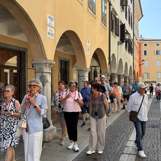 Eine Gruppe von Menschen, wahrscheinlich Touristen, läuft auf dem Bürgersteig einer Stadtstraße. Die Straße hat gewölbte Durchgänge, Gebäude und Straßenlaternen. Einige von ihnen tragen Sonnenbrillen, Armbänder und Uhren.
