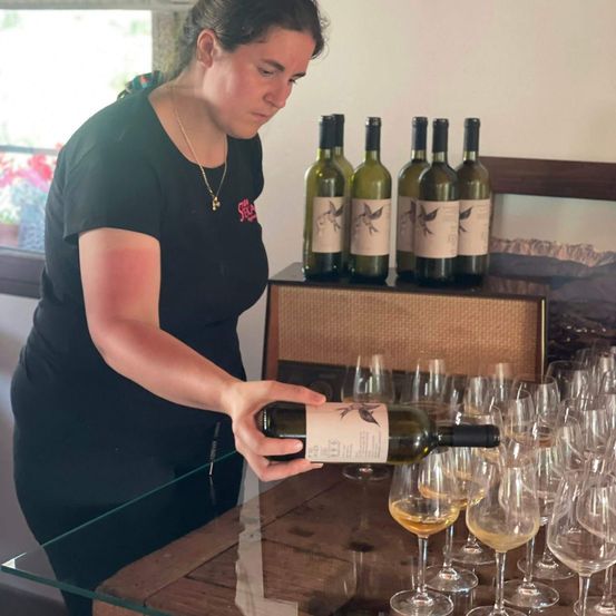 Eine Frau schenkt Wein aus einer Flasche in ein Weinglas, umgeben von anderen Weingläsern und Flaschen.