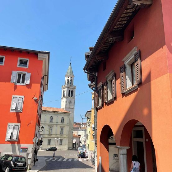 Eine Straßenszene in einer Stadt mit orangefarbenen Gebäuden und einem hohen Glockenturm. Eine Person geht in der Nähe eines Bogens, und Autos sind auf der Seite geparkt.