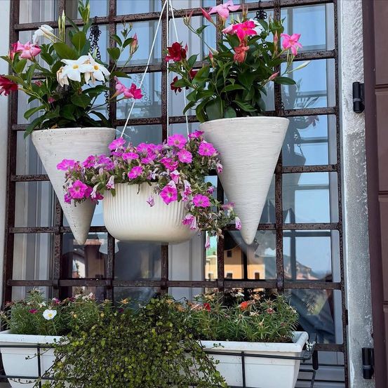 Drei weiße kegelförmige Pflanzgefäße mit bunten Blumen hängen an einem Fenster mit einem Metallgitter. Unten befinden sich zwei rechteckige weiße Pflanzgefäße mit grünen Pflanzen.