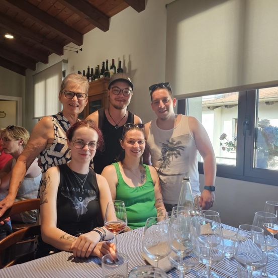 Eine Gruppe von Menschen, wahrscheinlich Familie, posiert für ein Foto in einem Essbereich mit Weingläsern, einem Tisch und einem Fenster, das ein Gebäude und Bäume draußen zeigt.