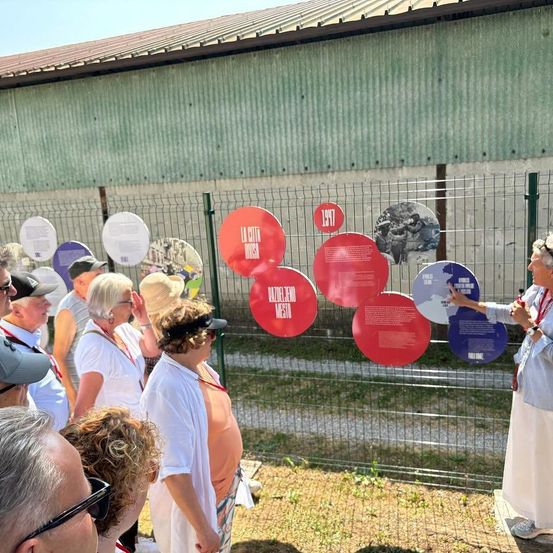 Touristen besuchen eine Gedenkstätte und betrachten Informationskreise an einem Zaun, wobei eine Person auf einen Kreis zeigt.