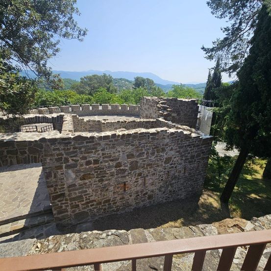 Eine steinerne Struktur mit einem zerstörten Turm steht zwischen Bäumen und bietet einen Panoramablick auf eine Berglandschaft unter einem klaren Himmel.