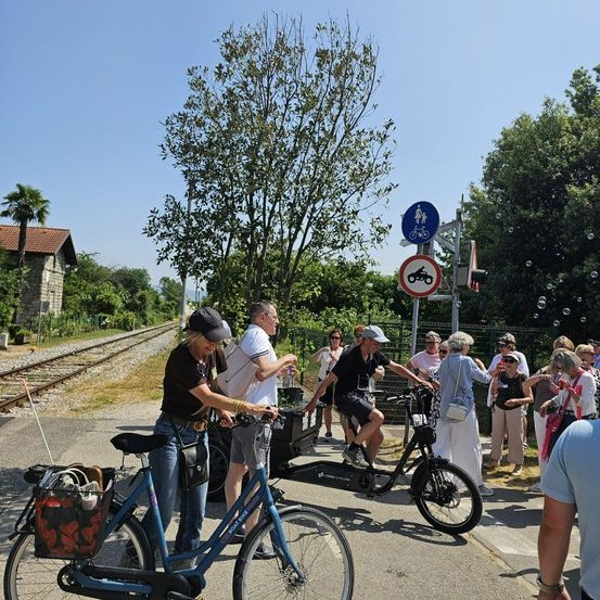 Eine Gruppe von Menschen auf Fahrrädern steht an einer Straßenecke. Eine Frau justiert ihr Fahrrad, während ein Mann mit ihr spricht. Ein Verkehrsschild befindet sich in der Nähe.
