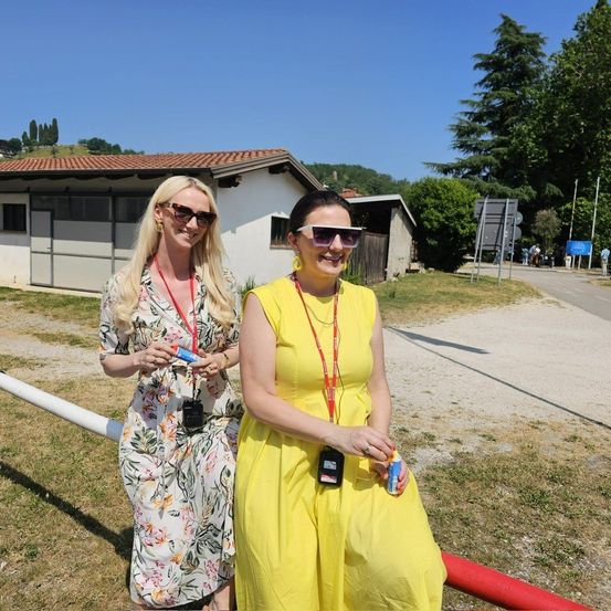 Zwei Frauen in Sommerkleidern und Sonnenbrillen stehen neben einem Pfahl. Eine hält eine weiße Tube, die andere eine blaue Dose. Hinter ihnen steht ein Gebäude mit rotem Dach. Bäume und Hügel sind in der Ferne.