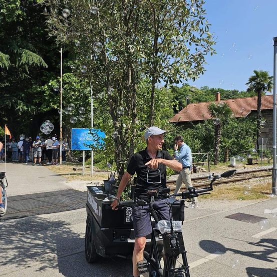 Ein Mann fährt mit einem Anhänger ein Fahrrad auf einer Straße, in der Nähe eines Hauses mit einem roten Dach. Menschen stehen hinter ihm und schauen zu. Bäume und Seifenblasen sind in der Luft.