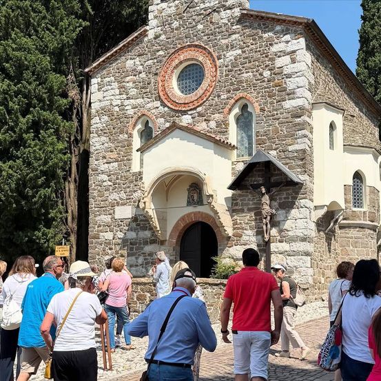 Eine Gruppe von Menschen steht vor einer alten Steinkirche mit einem Kruzifix an der Wand. Die Kirche hat ein rundes Fenster, einen gewölbten Eingang und ein Kreuz über dem Eingang.
