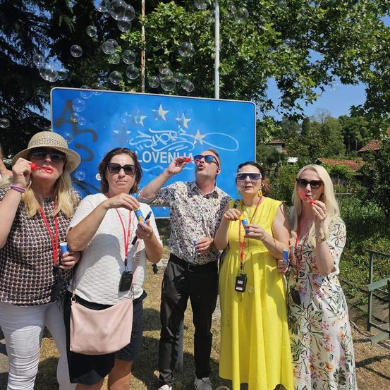 Fünf Personen stehen vor einem blauen Banner mit den Worten 'Eurovision' und 'Lovvenija' darauf. Sie tragen alle Sonnenbrillen und halten Seifenblasenmacher. Es schweben Seifenblasen in der Luft.