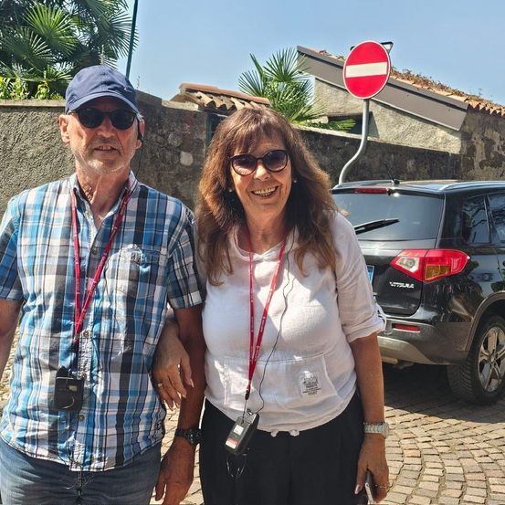 Ein älteres Paar steht lächelnd auf einer Kopfsteinpflasterstraße. Der Mann trägt ein kariertes Hemd und eine Kappe, und die Frau trägt ein weißes Oberteil und eine Sonnenbrille. Ein Auto ist hinter ihnen geparkt, mit einem roten Schild, das ein Verbot anzeigt.