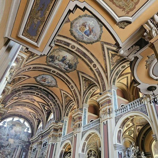Das Innere einer Kirche mit einer goldenen Decke, die religiöse Gemälde und aufwendige Verzierungen aufweist. Die Bögen und Säulen sind mit kunstvollen Designs geschmückt.