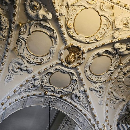 Eine reich verzierte Decke mit Goldakzenten zeigt Cherubs und Trompeten, einen großen zentralen Kreis umgeben von kleineren und kunstvollen Schnitzereien. Die Decke ist weiß mit goldenen dekorativen Elementen.