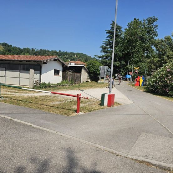 Ein Fahrradweg führt an einem weißen Haus mit rotem Dach vorbei. Ein Poller mit roten und weißen Streifen ist auf dem Weg positioniert. Bäume und Büsche umgeben die Szene.