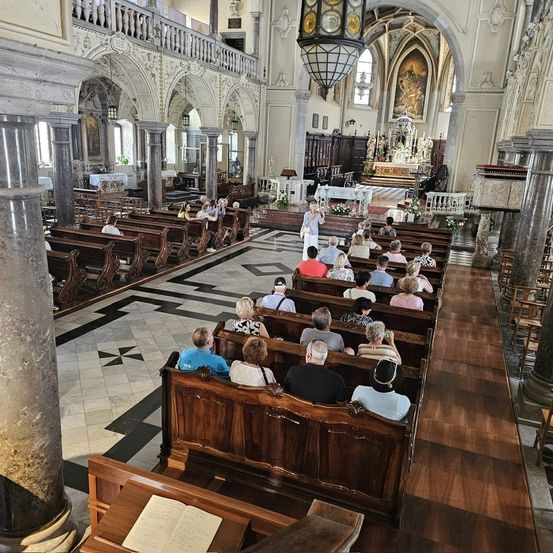 Im Inneren einer Kathedrale sitzen mehrere Menschen in Kirchenbänken, die zum Altar ausgerichtet sind, geschmückt mit Blumen und Kerzen. Eine Person steht am Altar und leitet wahrscheinlich einen Gottesdienst. Die Kathedrale zeichnet sich durch kunstvolle Säulen, komplizierte Bögen und eine hohe Decke aus.