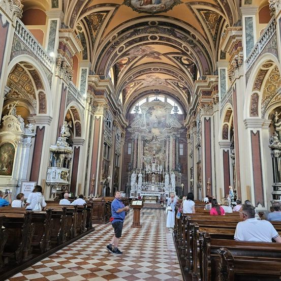 Ein reich verzierter Kircheninnenraum mit Menschen auf hölzernen Kirchenbänken. Ein Mann steht in der Nähe des Altars, andere schauen sich um. Die Kirche zeichnet sich durch aufwendige Bögen, Fresken und Statuen aus.