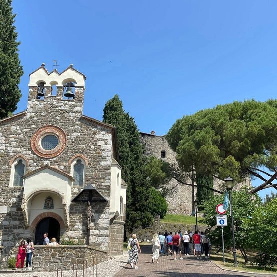Eine Steinkirche mit zwei Glocken im Turm. Eine Gruppe von Menschen geht davor.