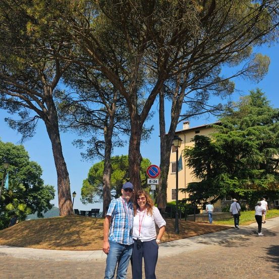Ein Mann und eine Frau stehen vor hohen Bäumen mit einem Schild in einem Stadtpark. Hinter ihnen gehen Menschen auf einem Kopfsteinpflasterweg, und ein Gebäude ist sichtbar.