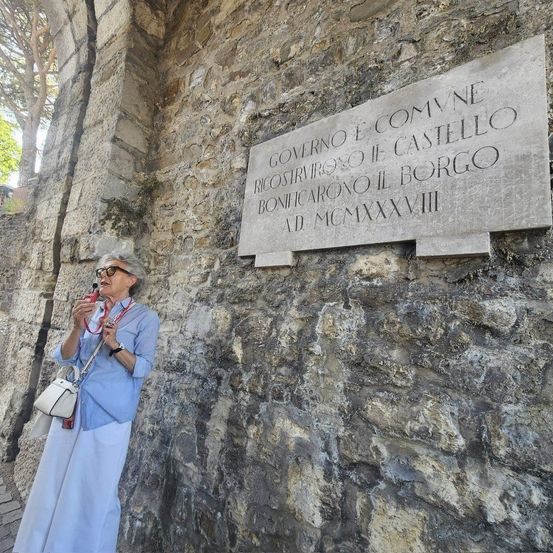 Eine Frau in einem blauen Outfit steht neben einer Steinmauer mit einer historischen Plakette. Die Plakette ist in Latein verfasst, das Jahr 1828 ist sichtbar. Sie hält ein Mikrofon, wahrscheinlich gibt sie eine Führung.