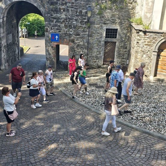 Touristen gehen durch einen Steinbogen auf einer Kopfsteinpflasterstraße, einige machen Fotos und andere unterhalten sich. Eine Lampe ist an einer Steinmauer montiert.