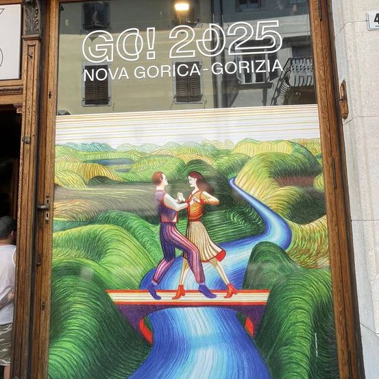 Ein Poster am Schaufenster eines Ladens zeigt ein Paar, das auf einer Brücke tanzt. Die Brücke überspannt einen Fluss in einem grünen Tal, mit den Worten 'GO! 2025' und 'NOVA GORICA-GORIZIA' oben. Das Poster hat einen lebendigen und farbenfrohen Stil.