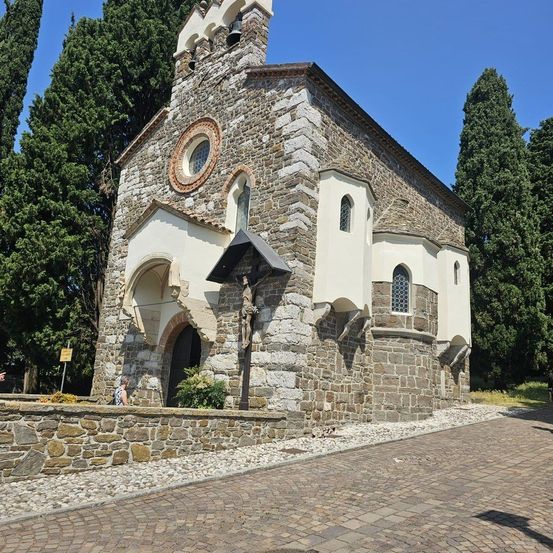 Eine kleine Steinkirche mit Glockenturm steht in einer ruhigen Umgebung. Sie hat ein rundes Fenster, einen Bogeneingang und eine Statue an der Wand. Bäume und ein gepflasterter Weg umgeben die Kirche.