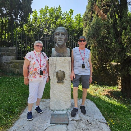 Zwei Personen stehen neben einem Büstendenkmal auf einem Steinsockel, tragen Sonnenbrillen und Freizeitkleidung. Sie befinden sich in einem Park mit grünem Rasen und Bäumen im Hintergrund.