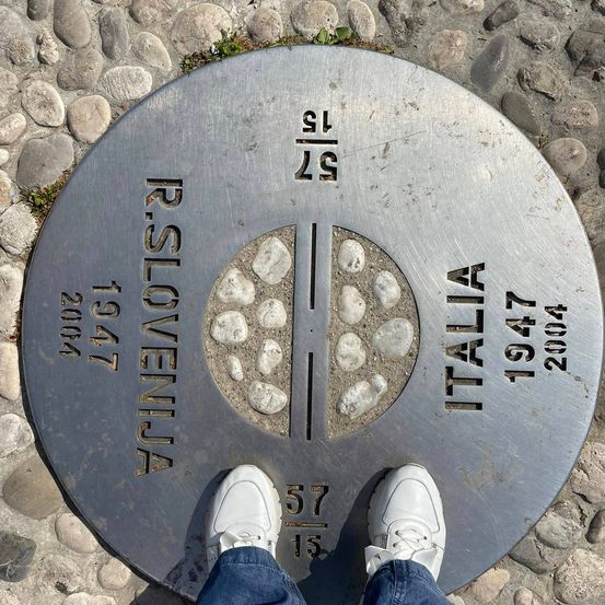 Eine Edelstahlplatte zeigt Italien und Slowenien mit Koordinaten 57 13 und 19 47, datiert 1947 und 2004. Eine Person steht darauf in weißen Turnschuhen.