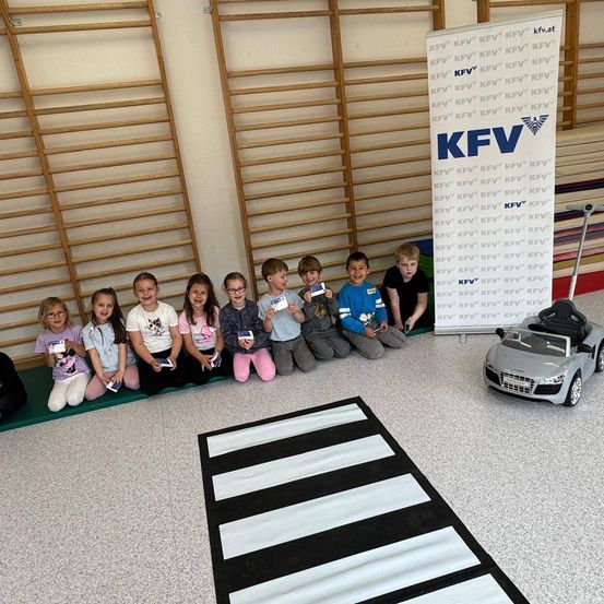 Eine Gruppe von Kindern sitzt auf einer grünen Matte in einem Raum und hält Urkunden. Hinter ihnen steht ein Schild mit 'KFV' und einem Logo auf einem Ständer. Ein Spielzeugauto steht rechts neben einer weiß-schwarz gestreiften Matte.
