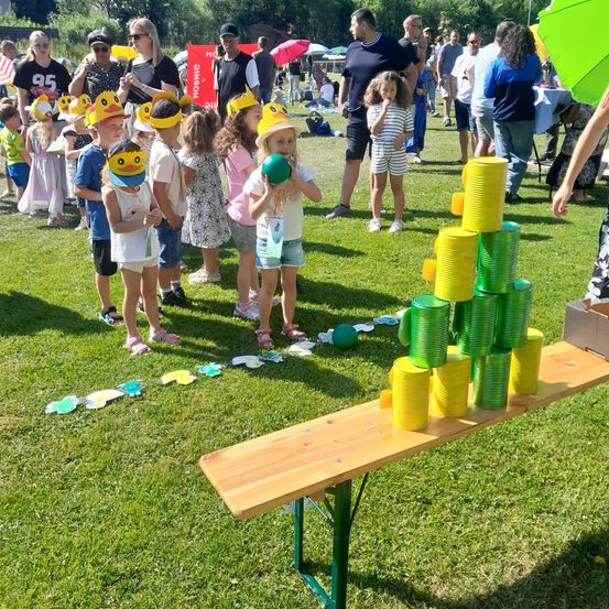 Eine Gruppe von Kindern mit gelben Hüten mit Schnäbeln spielt auf einem Grasfeld. Sie stehen in einer Reihe und halten Bälle, wahrscheinlich nehmen sie an einem Spiel teil. Dahinter sind Erwachsene und ein Tisch mit gestapelten Dosen.
