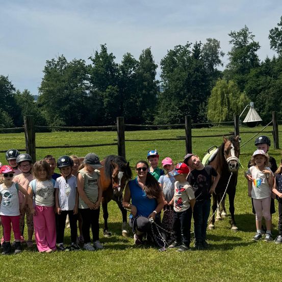 Eine Gruppe von Kindern und Erwachsenen versammelt sich auf einer Wiese mit zwei Pferden, trägt Helme und Freizeitkleidung. Die Umgebung ist im Freien mit einem Zaun und Bäumen im Hintergrund.