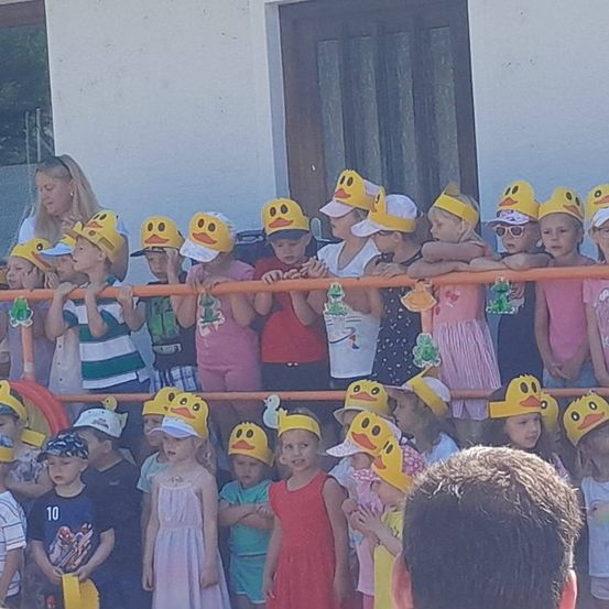 Eine Gruppe von Kindern in Hüten mit gelben Enten darauf stehen vor einem Gebäude. Sie schauen über eine Barriere, wobei eine Frau hinter ihnen steht.