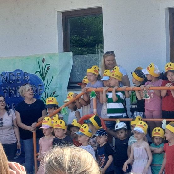 Eine Gruppe von Kindern mit Entenhüten steht vor einem Gebäude mit zwei Frauen. Eine Frau hält ein Banner mit Text. Die Kinder scheinen an einer Schulveranstaltung teilzunehmen.