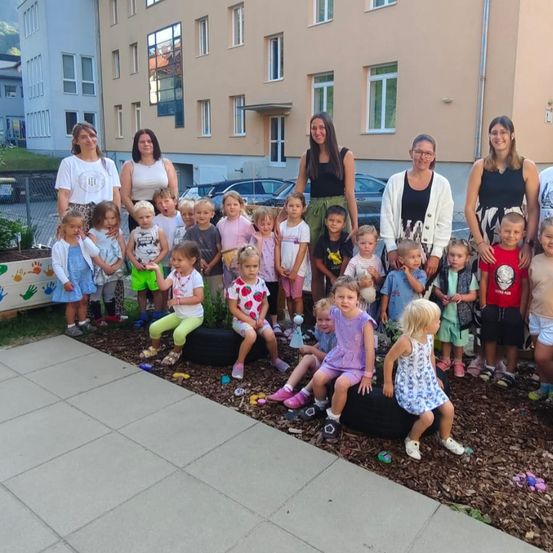 Eine Gruppe von Kindern und Erwachsenen posiert für ein Foto in einem Garten, einige sitzen auf Reifen. Hinter ihnen sind Gebäude mit Fenstern zu sehen.