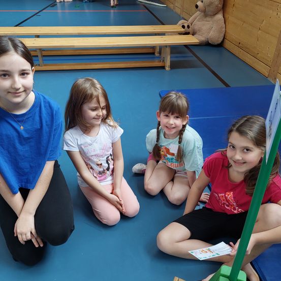 Vier junge Mädchen sitzen lächelnd auf einem blauen Boden. Sie sind in verschiedenfarbigen Shirts und Hosen gekleidet. Hinter ihnen ist eine Holzbank und ein brauner Teddybär zu sehen. Die Mädchen scheinen an einer Aktivität teilzunehmen oder für ein Foto zu posieren.