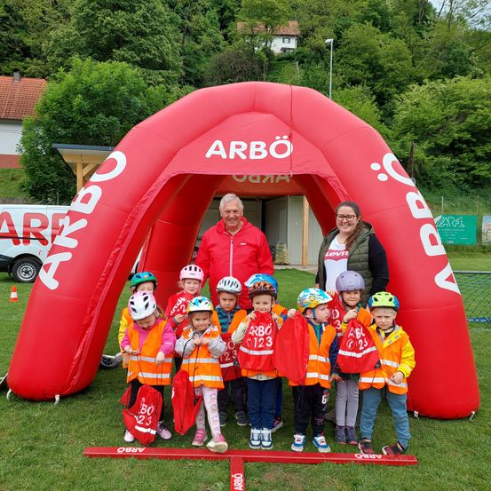 Eine Gruppe von Kindern und zwei Erwachsenen steht in einem roten ARBÖ-Bogen. Die Kinder tragen Sicherheitswesten und halten rote Fahnen. Die Erwachsenen lächeln. Dahinter ist ein Gebäude zu sehen, und es gibt Bäume und eine Straßenlaterne im Hintergrund.