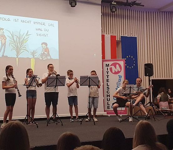 Eine Gruppe von Kindern spielt auf der Bühne Musikinstrumente. Ein Beamer zeigt einen deutschen Text. Zwei Erwachsene begleiten sie. Ein Publikum sitzt vor der Bühne.