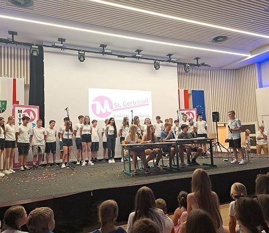 Eine Gruppe von Schülern steht auf einer Bühne mit einer Projektionsleinwand und einem Mikrofon, während andere an Tischen sitzen. Das Publikum beobachtet von vorne.