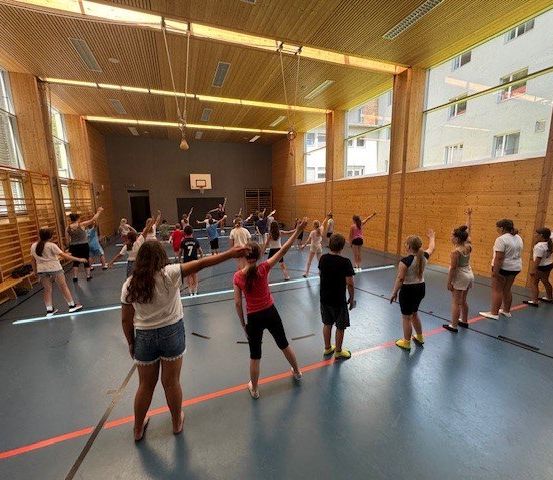 Eine Gruppe von Menschen übt Tanzbewegungen in einer Turnhalle mit Holzwänden und großen Fenstern. Sie stehen in Reihen, manche mit erhobenen Armen. Ausrüstung ist auf der linken Seite platziert.