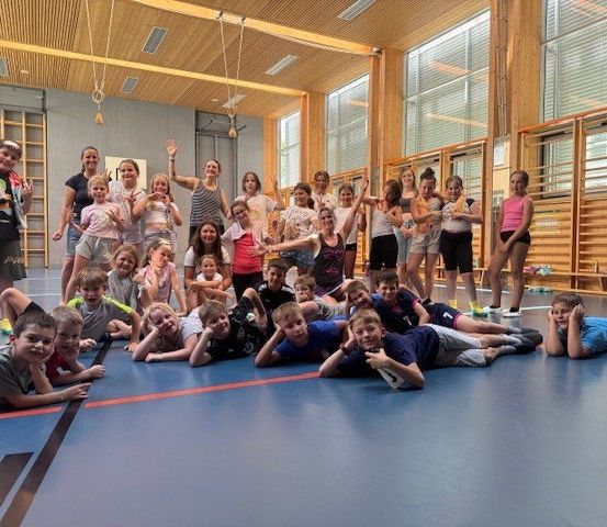 Eine Gruppe von Kindern und Jugendlichen versammelt sich in einer Turnhalle, einige sitzen auf dem Boden, andere stehen, wobei mehrere Erwachsene anwesend sind. Sie scheinen für ein Foto zu posieren.