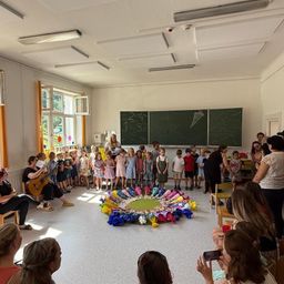 Eine Gruppe von Kindern und Erwachsenen versammelt sich in einem Klassenzimmer, einige sitzen und andere stehen um einen bunten Kreis auf dem Boden herum. Eine Frau sitzt mit einer Gitarre, und eine Tafel mit Zeichnungen ist im Hintergrund.