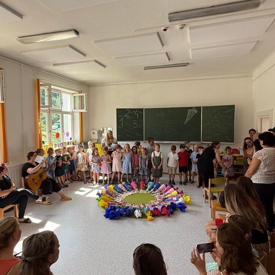 Eine Gruppe von Kindern und Erwachsenen versammelt sich in einem Klassenzimmer, einige sitzen und andere stehen um einen bunten Kreis auf dem Boden herum. Eine Frau sitzt mit einer Gitarre, und eine Tafel mit Zeichnungen ist im Hintergrund.