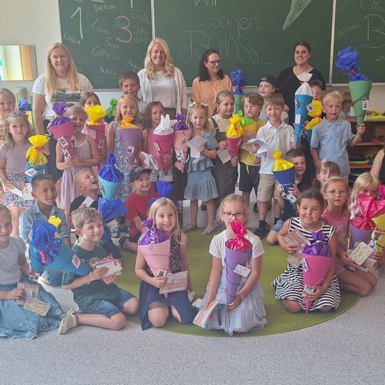 Eine Gruppe von Kindern in einem Klassenzimmer, die Blumensträuße halten, mit zwei Frauen, die hinter ihnen stehen. Die Tafel hinter ihnen hat Schrift darauf.