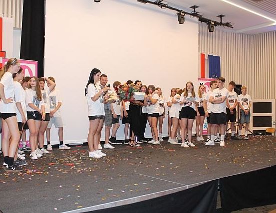 Eine Gruppe von Schülern steht auf der Bühne, hält Bücher und Blumen in den Händen, Konfetti verstreut darunter, ein Mann spricht in ein Mikrofon.