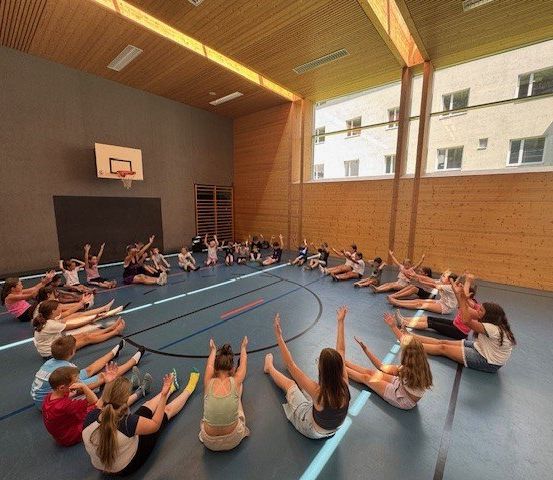 In einer Turnhalle sitzen eine Gruppe von Menschen im Kreis auf dem Boden und heben die Hände. An der Wand ist ein Basketballkorb angebracht.